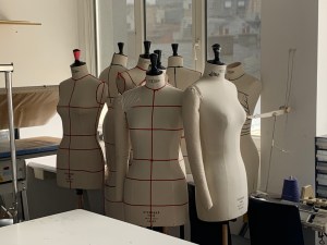 Photo of a cluster of white cloth mannequins, some with taped guide lines for clothing design, in a studio at the Paris College of art. In the foreground is a corner of a sewing table and in the background are various fashion design supplies. 