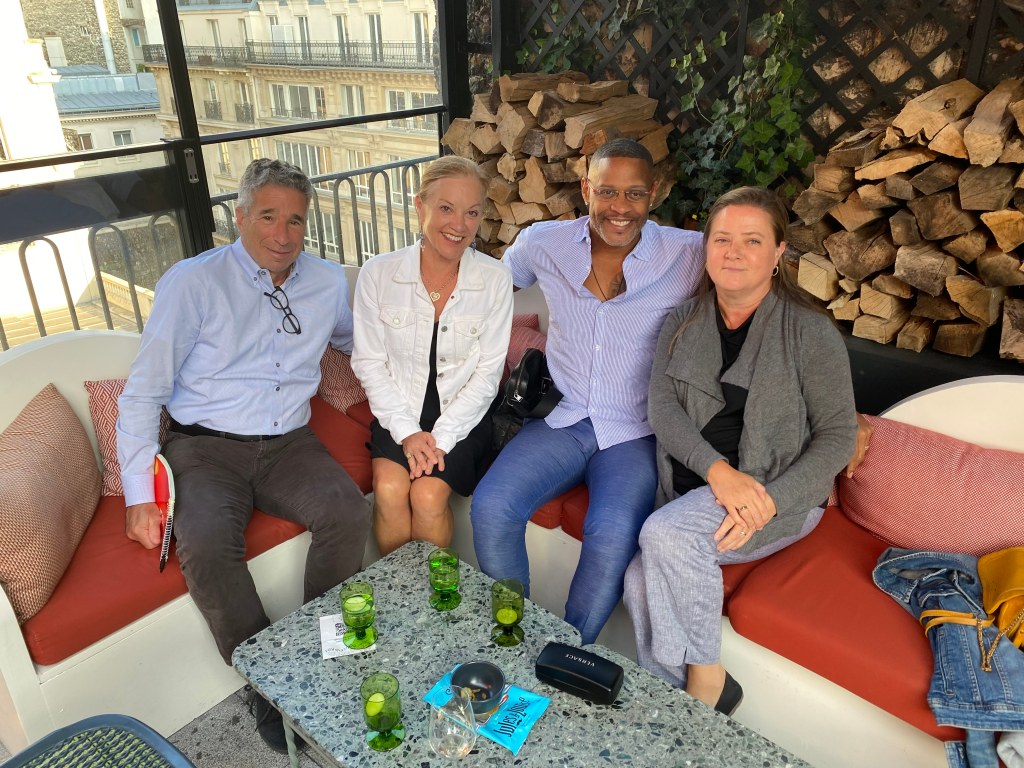 Photo at a rooftop restaurant. Photo shows, from left to right, Dr. Larry Schall from NECHE, his wife Betty Londergan,  Dr. Anthony Pinder, Vice President of International Affairs at Emerson College and President Linda Jarvin from the Paris College of Art, sitting on a corner couch with a table in front of them. Behind them is a beautiful window view of the city on the left side and a mix of wood and garden ivy on the right. 